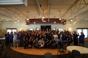 Large group of people indoors posing with fists raised in unity, around tables and chairs under bright ceiling lights in a hall