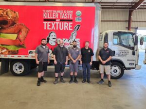 Five men stand in a warehouse in front of a red promotional truck trailer with a sandwich image and logos in the background.