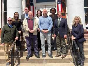 Group of nine adults standing on the steps of a brick building with white columns and American flags in the background.
