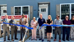 Ribbon-cutting ceremony in front of a blue house with a long blue ribbon and several officials holding the ribbon and scissors. - Participants include men in suits and a woman in a pink checkered suit, smiling for the photo.
