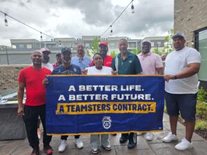 Group of diverse adults posing outdoors behind a blue banner that reads 'A BETTER LIFE. A BETTER FUTURE. A TEAMSTERS CONTRACT'