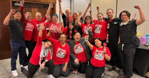 Large group of nurses inside, mostly wearing red, smiling, with raised fists