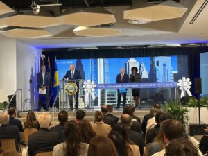 Speakers at a podium during a press conference on a stage with a blue cityscape backdrop and an audience watching.