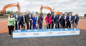 Group of professionals in business attire with ceremonial shovels at a groundbreaking ceremony on a construction site, with a city skyline in the background and heavy machinery nearby.