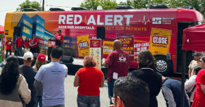 People speak at podium in front of Red Alert Tour bus, crowd watching