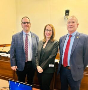 RI-Warehouse-Worker-Protection-Act-Lobbying-LRjpg - USA Works - USA Works Three professionals pose for a photo in a formal meeting room; woman in black blazer center, flanked by two men in suits and ties.