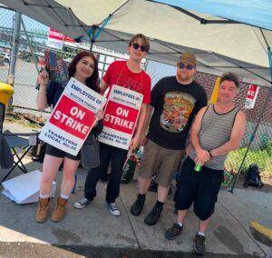 Boston-Towjpg - USA Works - USA Works Four people stand under a canopy at a protest, holding red-and-white 'ON STRIKE' signs for Boston Towing and Teamsters Local No. 25, with a chain-link fence in the background.