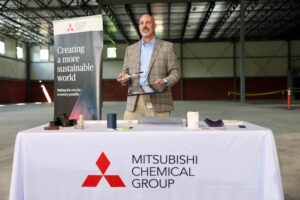 Mitsubishi-Chemical-Advanced-Materials-to-Expand-Operations-1jpg - USA Works - USA Works Man in a tan plaid blazer at a table with scientific props, standing beside a Mitsubishi Chemical Group banner in a large showroom or conference hall.