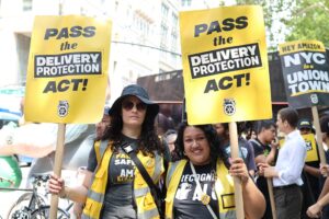 DPA-Picket-2-scaledjpeg - USA Works - USA Works Protesters hold large yellow signs reading 'PASS the DELIVERY PROTECTION ACT' at an urban street demonstration.