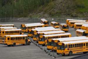 Parking lot filled with numerous yellow school buses, arranged in rows beside a chain-link fence and hillside trees.